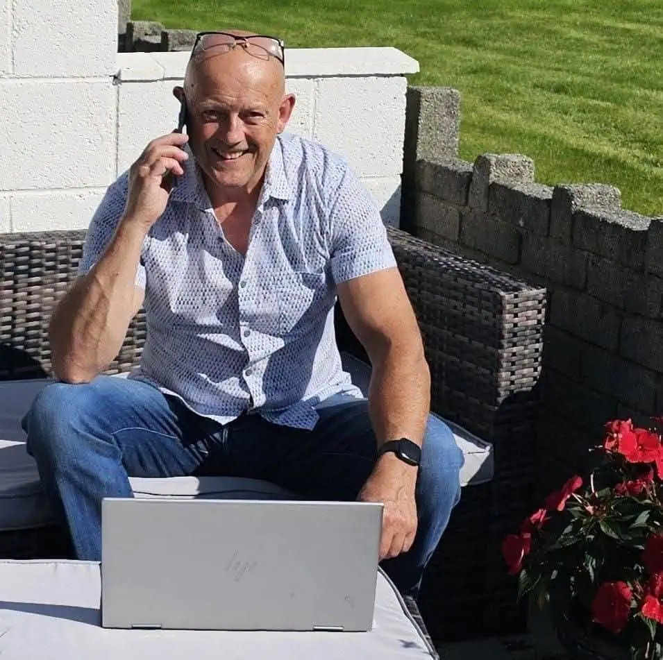 Smiling man seated outdoors on a couch, talking on a cellphone with a laptop in front of him.